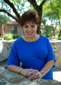 A woman in blue shirt sitting on ledge near trees.